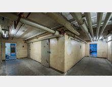 Kellerraum mit cremefarbenen Wänden und freiliegenden Rohren an der Decke. Zwei schwere Metalltüren und helle Beleuchtung dominieren den industriellen Raum, der mehrere Durchgänge und einen gefliesten Boden zeigt.