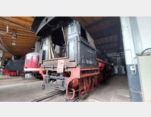 Historische Dampflok mit der Nummer "50 3682-7" in einer offenen, hölzernen Lokhalle. Im Vordergrund sind Details des Fahrwerks und Führerhauses zu sehen, im Hintergrund weitere historische Züge.