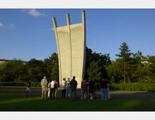 Mehrere Personen stehen vor einem hohen, dreiteiligen Betonmonument, bekannt als das Luftbrückendenkmal, in einem Park mit grünen Rasenflächen und Bäumen im Hintergrund. Der Himmel ist blau mit wenigen Wolken.