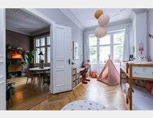 Wohnbereich mit zwei Räumen: im Vordergrund ein helles Kinderzimmer mit Holzboden, Hochbett, Spielzelt und Schreibtisch vor großen Fenstern; im Hintergrund ein Esszimmer mit Esstisch, Stühlen, Pflanzen und Bücherregal, sichtbar durch eine offene weiße Doppeltür.