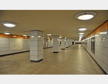 Unterirdischer Fußgängertunnel mit weißen, gefliesten Wänden, rechteckigen Säulen und mehreren an der Decke montierten runden Leuchten. Im Hintergrund führen Treppen hinauf, und an den Wänden befinden sich Informations- und Werbetafeln.