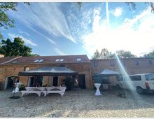 Hof mit einem historischen Backsteingebäude und gepflastertem Boden, ausgestattet mit Pavillons, dekorierten Stehtischen und einem geparkten Kleinbus. Im Hintergrund sind Bäume und ein strahlender Himmel mit Wolken zu sehen.