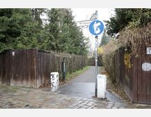 Ein schmaler Fußweg zwischen Holzzäunen und Hecken, mit einem Schild, das auf eine Fußgängerzone hinweist. Im Hintergrund sind hohe Bäume und vereinzelte Sträucher, der Weg ist gepflastert und nass.