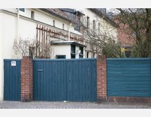 Hinterhofansicht mit einer cremefarbenen Gebäudeaußenwand, blauen Holzwänden und roten Backsteinsäulen, ergänzt durch karge Sträucher und Bäume. Im Hintergrund sind Ziegeldächer sowie ein Weihnachtsstern an einem Baum zu erkennen.