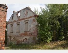 Verfallenes, zweistöckiges Gebäude aus Ziegelstein mit beschädigter Fassade und zugemauerten Fenstern, umgeben von dichter Vegetation und Bäumen; im Vordergrund ein schmaler Streifen Gras und ein Mauerrest aus roten Ziegeln.