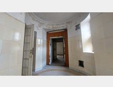 Rundförmiger Innenraum mit beigefarbenen Fliesenwänden und abgeblätterter Farbe an der Decke, einer offenen Tür mit Holzrahmen, die in einen weiteren Raum mit weißen Backsteinwänden führt. Rechts ein kleines Fenster und eine offene Lüftungsöffnung.