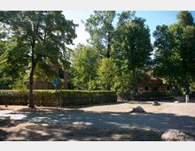 Parkähnliche Umgebung mit gepflastertem Weg, großen Laubbäumen und zwei Gebäuden im Hintergrund, eines aus rotem Backstein und eines im Fachwerkstil. Im Vordergrund ein Verkehrsschild für Radfahrer sowie ein mit Hecken eingefasster Zaun.