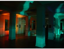 Ein Innenraum mit hohen weißen Säulen, farblich beleuchtet in Orange- und Grüntönen. Im Hintergrund sind große Fenster mit Rundbögen, Treppengeländer und Wandelemente zu sehen.