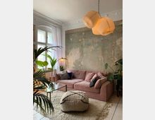 Wohnzimmer mit einer rosa Couch vor einer freigelegten, teils abgeblätterten Wand in Betonoptik. Der Raum ist mit Pflanzen, einer Hängelampe mit papierartigen Lampenschirmen, einem flauschigen Teppich und einem Glas-Couchtisch eingerichtet. Große Fenster mit weißen Vorhängen lassen Tageslicht herein.