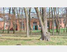 Backstein-Gebäude mit großen Fenstern, umgeben von Bäumen auf einer Grasfläche. Im Vordergrund stehen kahle Bäume, während das Gebäude im Hintergrund eine geöffnete grüne Tür zeigt.