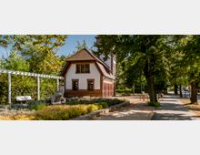 Kleines weißes Gebäude mit rotem Ziegeldach und Fassadenelementen aus Backstein, umgeben von einem Garten mit niedrigen Pflanzen und einer Pergola mit weißer Bank; rechts daneben ein gepflasterter Fußweg, gesäumt von großen Bäumen.