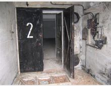 Metallene Tür mit der großen Ziffer "2", die teilweise geöffnet ist, führt in einen leeren, dunklen Raum. Die Umgebung zeigt eine verwitterte Betonwand mit freiliegenden Kabeln und alten elektrischen Schaltanlagen.