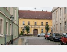 Eine kleine gepflasterte Straße mit geparkten Autos führt zu einem gelben, zweistöckigen Rathausgebäude mit roten Dachziegeln. Vor dem Gebäude befinden sich Laternen, Bäume und Blumen, umgeben von angrenzenden Gebäuden in Grün- und Beigetönen.