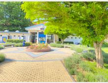 Eingangsbereich eines kleinen Gebäudekomplexes mit weiß-blauer Fassade und einem Uhrenturm. Im Vordergrund eine gepflasterte Fläche mit Blumenbeet, Sitzbänken und umgeben von Bäumen und Sträuchern.