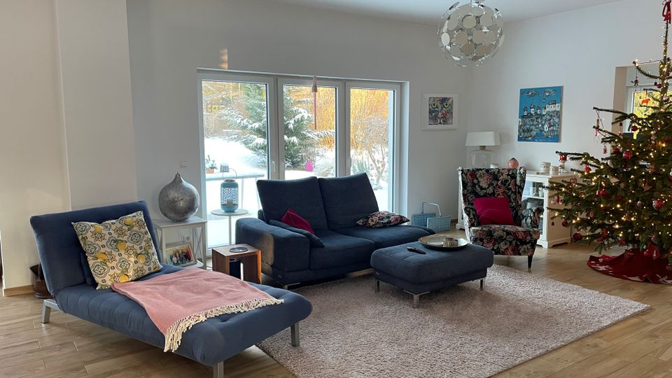 Helles Wohnzimmer mit großen Glas-Schiebetüren zur verschneiten Terrasse, Holzfußboden und hellem Teppich. Im Raum stehen ein dunkelblaues Sofa mit Hocker, eine Liege, ein gemusterter Sessel, ein Sideboard sowie ein geschmückter Weihnachtsbaum und moderne Deckenleuchte.