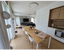 Moderne Wohnküche mit offenem Wohnbereich: Ein heller Raum mit Holzboden und weißen Möbeln, einer Küchenzeile mit Holzverkleidung, einem Esstisch mit weißen Stühlen und einem angrenzenden Wohnzimmer mit großem Fernseher und Sofas. Große Fensterfronten mit weißen Vorhängen lassen viel Tageslicht einfallen.