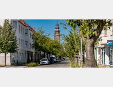 Straßenszene in einer Wohngegend mit mehrstöckigen Gebäuden auf beiden Seiten, teils mit Geschäften im Erdgeschoss, und Bäumen, die die Straße säumen. Im Hintergrund ragt ein historischer Turm mit einer Uhr in die Höhe.