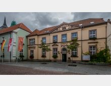Zweistöckiges, historisch gestaltetes Gebäude mit heller Fassade und roten Dachziegeln. Vor dem Gebäude befinden sich drei Fahnenmasten, kleine Bäume und Fahrräder; im Hintergrund ragt der Turm einer Kirche empor.