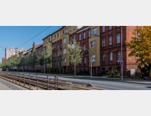 Straßenansicht mit mehrstöckigen Wohngebäuden aus Ziegelstein und Putz, teils in Rot und Gelb. Die Straße verläuft parallel zu Straßenbahnschienen, gesäumt von jungen Bäumen und Straßenlaternen.