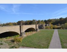 Eine alte Betonbrücke mit mehreren Bögen, die teilweise beschädigt ist, erstreckt sich über eine grüne Wiese. Im Hintergrund sind Bäume mit herbstlichem Laub und ein klarer blauer Himmel zu sehen; ein schmaler Gehweg führt am rechten Bildrand entlang.