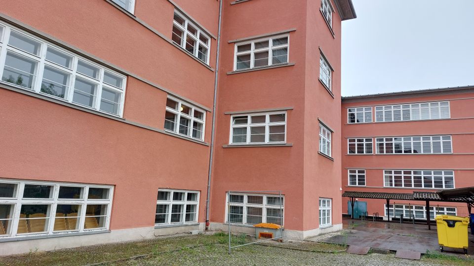 Ein mehrstöckiges Gebäude mit rosafarbenem Putz und weißen rechteckigen Fenstern. Der Hofbereich ist teilweise gepflastert und beinhaltet einen gelben Müllcontainer sowie ein Vordach aus Metall und Wellblech.