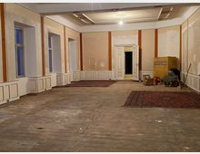 Großer, weitgehend leerer Saal in einem Altbau mit hohen Fenstern, Wandvertäfelung und dekorierter Kassettendecke; der Holzboden ist abgenutzt und zwei Teppiche liegen aus. Im Hintergrund steht eine offene Doppeltür, rechts sind Renovierungsmaterialien und ein Kachelofen zu sehen.