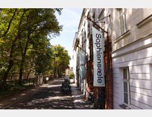 Straßenansicht einer ruhigen, von hohen Bäumen beschatteten Allee mit Gehweg, parkenden Fahrrädern und einem schmiedeeisernen Zaun entlang einer Grünanlage. Rechts stehen helle mehrstöckige Altbaufassaden mit einem roten Backsteineingang und einem großen vertikalen Banner mit der Aufschrift „SophienSaele“.