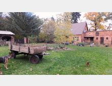 Hof mit Backsteingebäuden im ländlichen Stil, umgeben von viel Grün. Im Vordergrund stehen ein hölzerner Anhänger, ein Hund und ein Huhn auf einer Wiese; im Hintergrund befinden sich Bäume und Ziegelmauern.