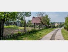 Eine ländliche Straße mit Betonplatten, gesäumt von Wiesen und Zäunen. Rechts neben der Straße stehen Häuser mit roten und gelben Dächern, während im linken Bereich ein eingezäuntes Grundstück mit einem alten Schuppen und Bäumen sichtbar ist.