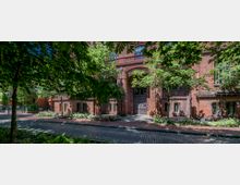 Historisches Backsteingebäude mit verzierten Metalltüren, flankiert von kleinen Fenstern und umrahmt von Bäumen und Grünflächen; im Vordergrund ein gepflasterter Gehweg und eine Straße.