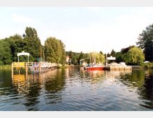 Eine Fluss- oder Seeuferlandschaft mit einem Steg und einem gelb beschrifteten Schild "Restaurant" auf der linken Seite. Rechts liegen mehrere Segelboote und Motorboote an einem kleinen Hafen, umgeben von Bäumen und Grünflächen.