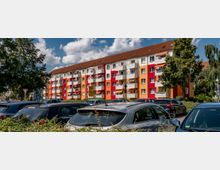 Mehrstöckiges Wohngebäude mit bunten Fassaden in Rot-, Orange- und Gelbtönen, ergänzt durch weiße Balkone, davor geparkte Autos und umgeben von Bäumen und Vegetation. Der Himmel ist teils bewölkt, und Verkehrsschilder sind am Straßenrand zu sehen.