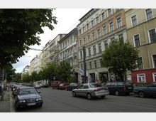 Straßenansicht eines Wohnviertels mit mehrstöckigen Altbauten, einigen mit buntem Putz und Graffiti, entlang einer Straße mit parkenden Autos und Straßenbahnschienen. Im Vordergrund befinden sich Bäume, die den Gehweg säumen.