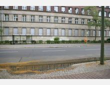 Straßenszene mit einem mehrstöckigen, beige-gelben Gebäude mit zahlreichen Fenstern im Hintergrund. Im Vordergrund befinden sich eine Bushaltestelle, nasse Pflastersteine mit einer Pfütze, eine Straße mit Fahrbahnmarkierungen und ein orangefarbener Briefkasten an einem Pfahl.