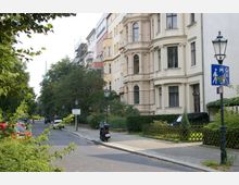 Straßenszene in einer Wohngegend mit Altbauten, verzierten Fassaden und großen Fenstern. Links der Straße parken Autos, rechts befinden sich Grünflächen mit Zäunen, sowie ein Gehweg und Straßenlaternen.