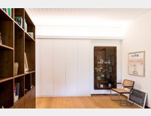 Ein schlichter Innenraum mit Holzboden, einem großen Bücherregal links, einer weißen Wand mit eingebauter Schiebetür in der Mitte und einer verglasten Vitrine rechts neben einem gerahmten Kunstwerk. Eine Stuhl-Sitzgruppe mit hellem Geflecht befindet sich am rechten Bildrand.