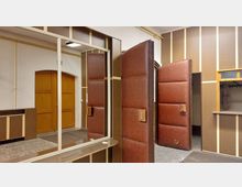 Innenansicht eines Raumes mit holzvertäfelten Wänden und gepolsterten braunen Türen. Zu sehen sind eine Spiegelwand, eine Garderobe mit Kleiderhaken und eine große Holztür im Hintergrund.