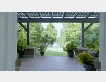 Überdachte Veranda mit dunklen Holzträgern und Ziegelpfeilern, umgeben von großen grünen Pflanzen in Töpfen, mit Blick auf einen gepflegten Garten und dahinterliegendem Wasser im Hintergrund.