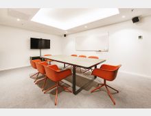 Konferenzraum mit einem rechteckigen Tisch, umgeben von acht orangefarbenen Stühlen auf Teppichboden. An der weißen Wand befinden sich ein Flachbildfernseher und ein Whiteboard.