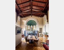 Innenraum einer umgestalteten Kirche mit hölzernem Gewölbedach, großen Rundbogenfenstern und einem zentral platzierten Buddha-Bildnis. Im Vordergrund steht ein rustikaler Holztisch mit Stühlen auf einem Parkettboden, umgeben von dekorativen Elementen und Blumenarrangements.