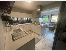 Moderne Küche mit weißer Ausstattung und einer Küchentheke in der Mitte, darüber eine silberne Dunstabzugshaube. Die Rückwand zeigt ein großformatiges Waldmotiv. Im Hintergrund befinden sich ein Essbereich mit großen Fenstern und Blick auf einen Garten.