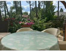Terrasse oder Garten-Sitzplatz mit rundem Tisch (hellgrüne Tischdecke) und Korbstühlen, umgeben von dichtem Grün und blühenden Sträuchern. Im Hintergrund stehen hohe Bäume, ein Metall-Pavillonrahmen und Dächer von Wohnhäusern unter blauem Himmel.