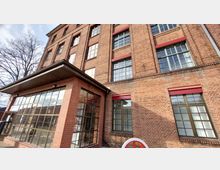 Backsteinfassade eines historischen Industriegebäudes mit großen, rechteckigen Fenstern und roten Fensterrahmen. Im Vordergrund ein moderner Anbau mit Glasfront und einem Vordach; im rechten unteren Eck ein Detail eines rot-weißen Schwungrades.