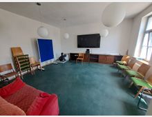 Ein einfacher, heller Gemeinschaftsraum mit grünem Teppichboden, mehreren Stühlen entlang der Wände, einer roten Couch im Vordergrund und einer großen schwarzen Wandtafel mit einem darunterliegenden Holzmöbel an der Stirnseite des Raumes. Drei große, runde Hängelampen hängen von der Decke, und durch ein Fenster auf der rechten Seite fällt Tageslicht ein.