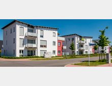 Mehrfamilienhäuser in einer Wohnsiedlung mit weißer und roter Fassade, Balkonen und Flachdächern; umgeben von einer ruhigen Straße, Gehwegen, jungen Bäumen und einer Straßenecke mit einem Wegweiser.