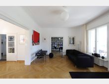 Wohnzimmer mit hellem Holzboden und weißen Wänden. Ein schwarzes Sofa, moderne Stühle, ein abstraktes Kunstwerk an der Wand und große Fenster prägen den Raum; im Hintergrund ist ein Bücherregal in einem angrenzenden Zimmer sichtbar.