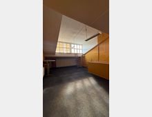 Leerer Innenraum mit schräger Decke, hohen Fenstern an der Stirnseite und hängenden Leuchtstofflampen. An der rechten Seite steht ein einfacher Tresen, daneben führt eine Leiter zu einer erhöhten Ablage unter den Fenstern.