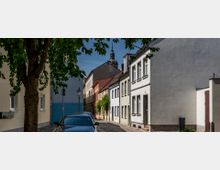 Eine enge Kopfsteinpflasterstraße in einer Altstadt mit zweistöckigen Häusern in verschiedenen Pastellfarben, rechts flankiert von einem großen Baum. Im Hintergrund ist ein Kirchturm mit Uhr und Spitze zu sehen.