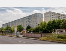 Straßenszene mit mehrstöckigen Plattenbau-Wohngebäuden im Hintergrund, umgeben von Bäumen. Im Vordergrund befinden sich eine Bushaltestelle mit Unterstand und Hinweisschildern sowie ein Kiosk.
