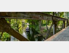 Ein schmaler, von Bäumen gesäumter Kanal, durch den ein Boot mit mehreren Personen fährt, aufgenommen durch die Struktur einer rostigen Metallbrücke im Vordergrund. Die Umgebung wirkt grün und idyllisch mit dichtem Bewuchs an den Ufern.
